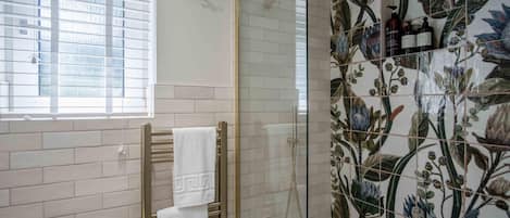 A bathroom with white tiled walls, a shower with a glass door, and a window with blinds.