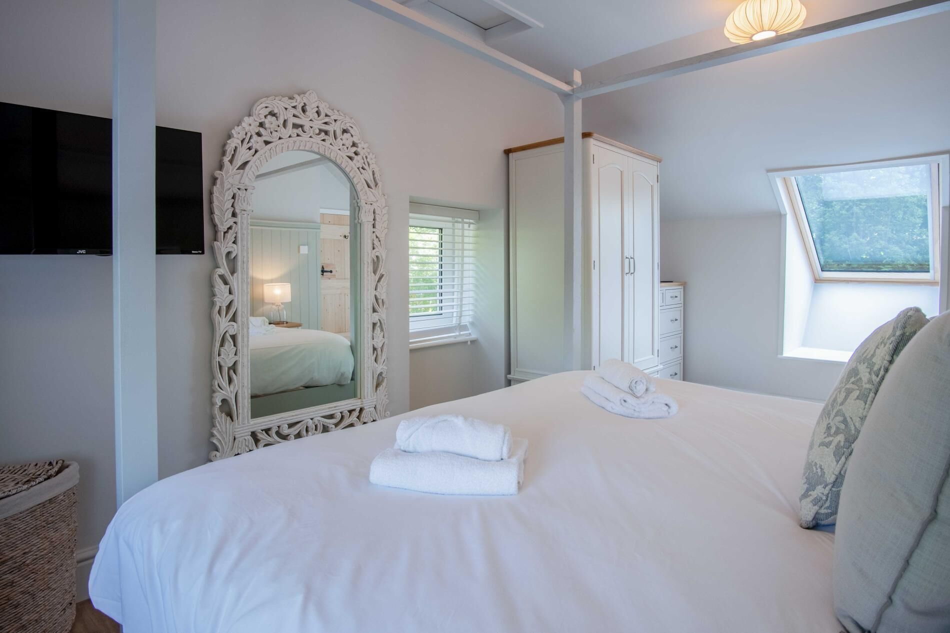 Une chambre à coucher avec un grand miroir décoratif, un lit recouvert de linge blanc, une armoire et un téléviseur fixé au mur.