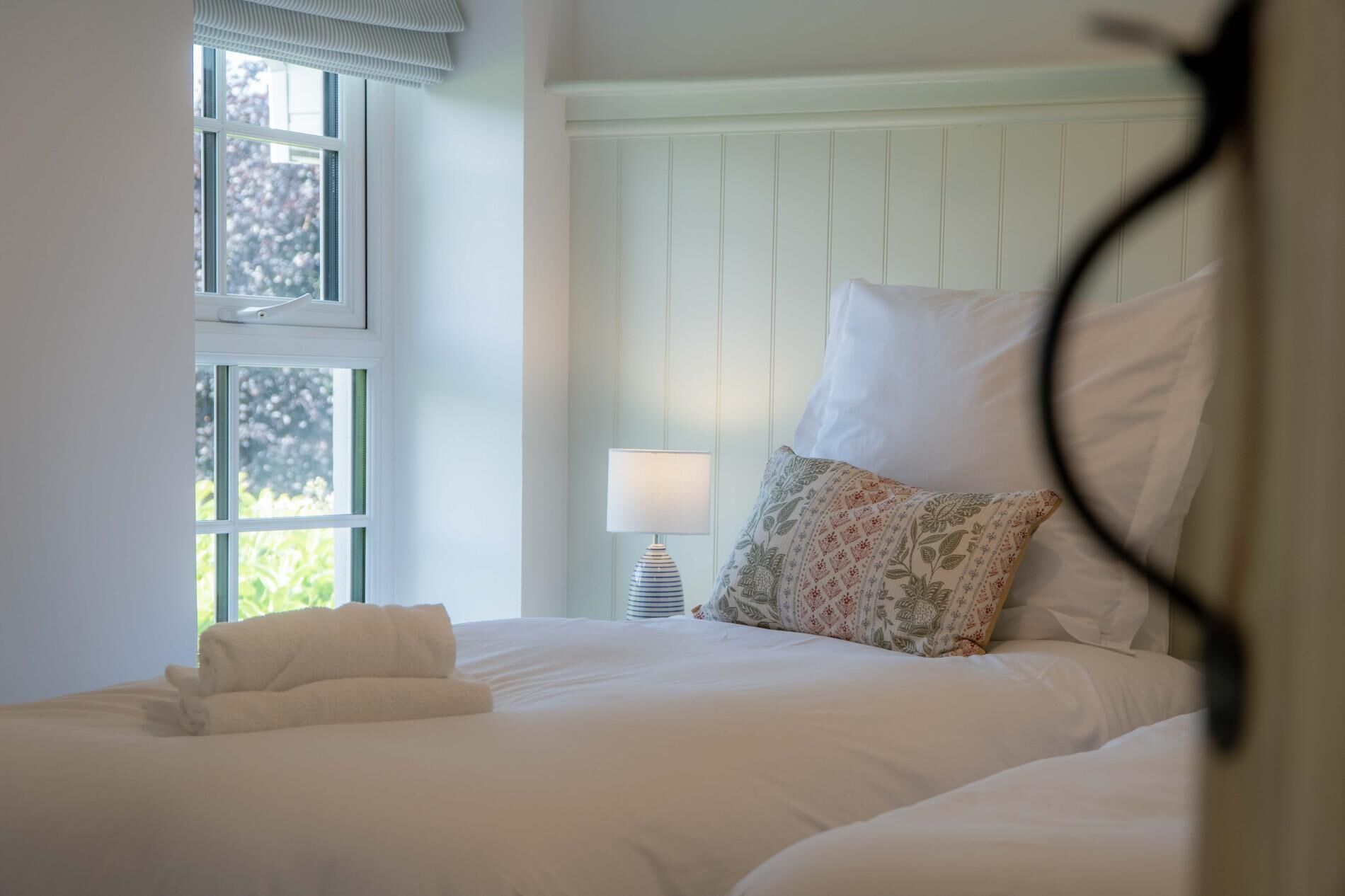 A neatly made bed with white linens and a floral pillow, a bedside lamp, and a window with a view of trees.