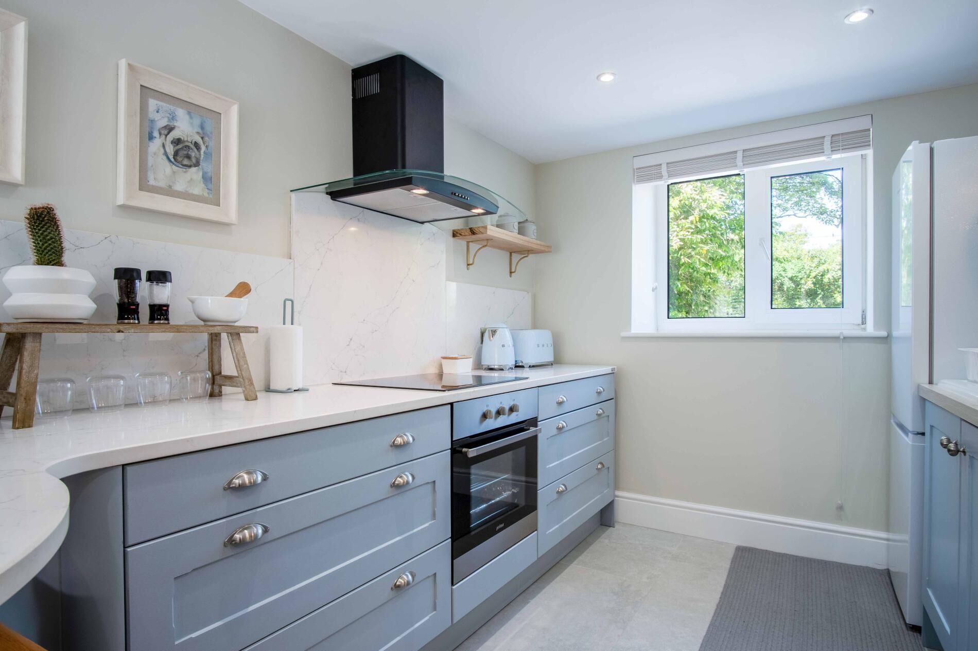 A modern kitchen with grey cabinets, a built-in oven, a black range hood, and a window with a view of greenery.