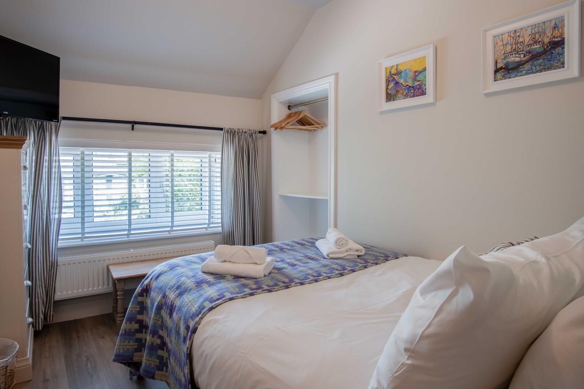A bedroom with a bed, a window with blinds, a radiator, a hanging coat rack, and two framed artworks on the wall.