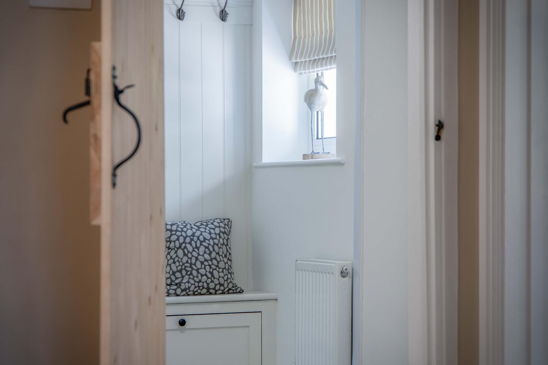 Ein Flur mit Heizkörper, einem Fenster mit Streifenrollo und einem Kissen mit Muster auf einem weißen Schrank.