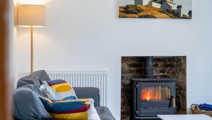 A living room with a grey sofa, colorful pillows, a white table, and a fireplace with a visible fire. - Fern Bank - 5 Bedroom House - Horton (Swansea)