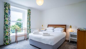 A neatly arranged bedroom with a large bed, two bedside tables, a wooden nightstand, a window with curtains, and a view of greenery outside. - Fern Bank - 5 Bedroom House - Horton (Swansea)