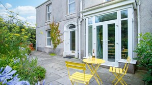 A two-story house with a patio area featuring a yellow table and chairs, a bottle of wine, and a variety of plants and flowers. - Fern Bank - 5 Bedroom House - Horton (Swansea)