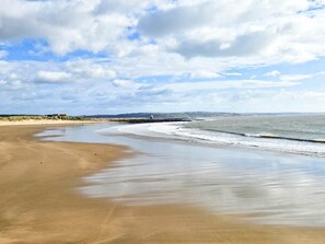 Ampia spiaggia sabbiosa con onde calme, cielo sereno e una costa lontana con edifici.