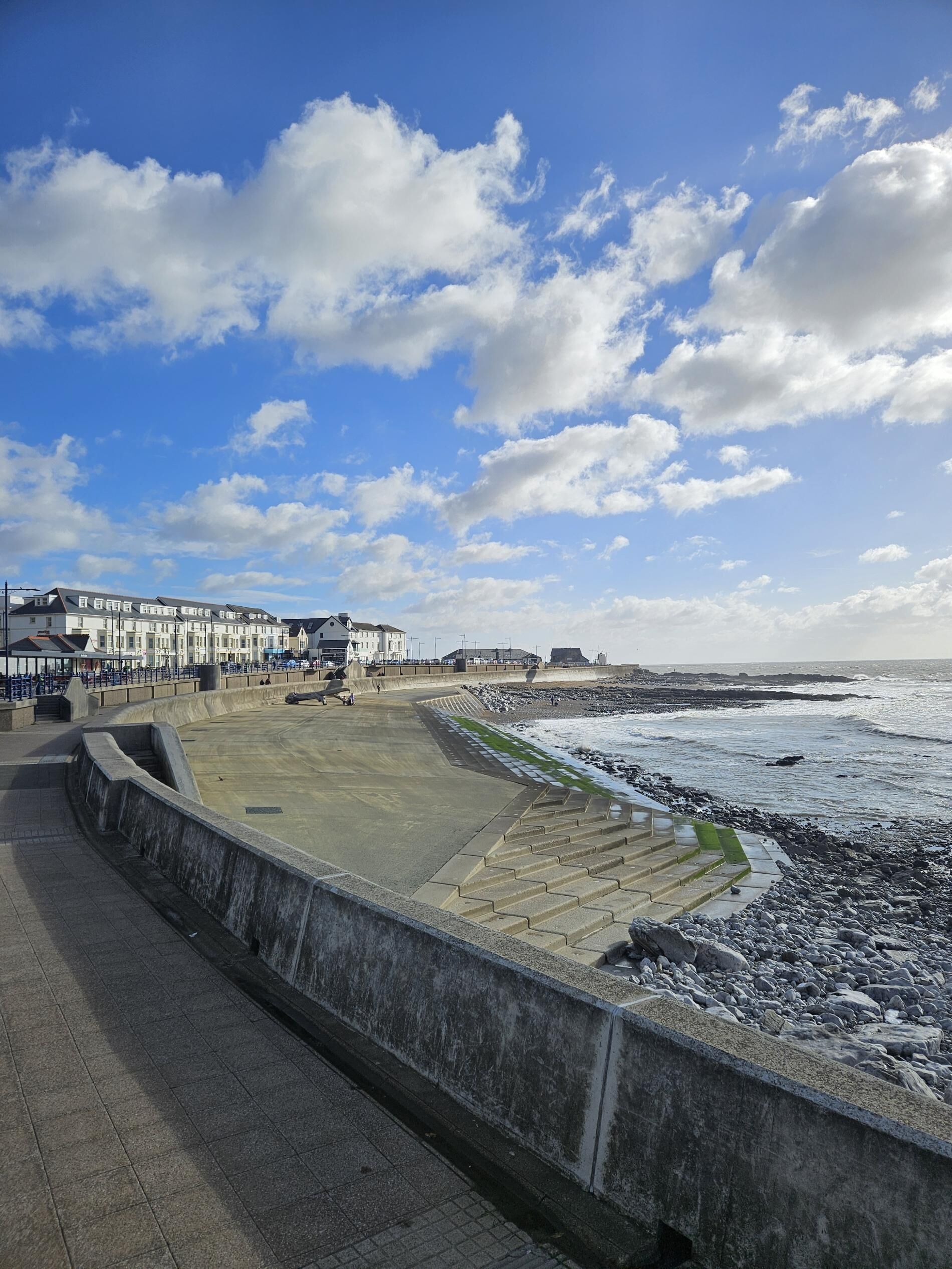 Un passeig marítim amb una passarel·la de formigó, un mur de pedra i vista al mar i edificis al fons.