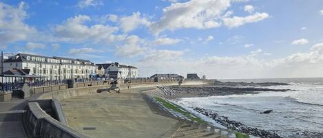 A coastal promenade with a concrete walkway, a stone wall, and a view of the sea and buildings in the background.