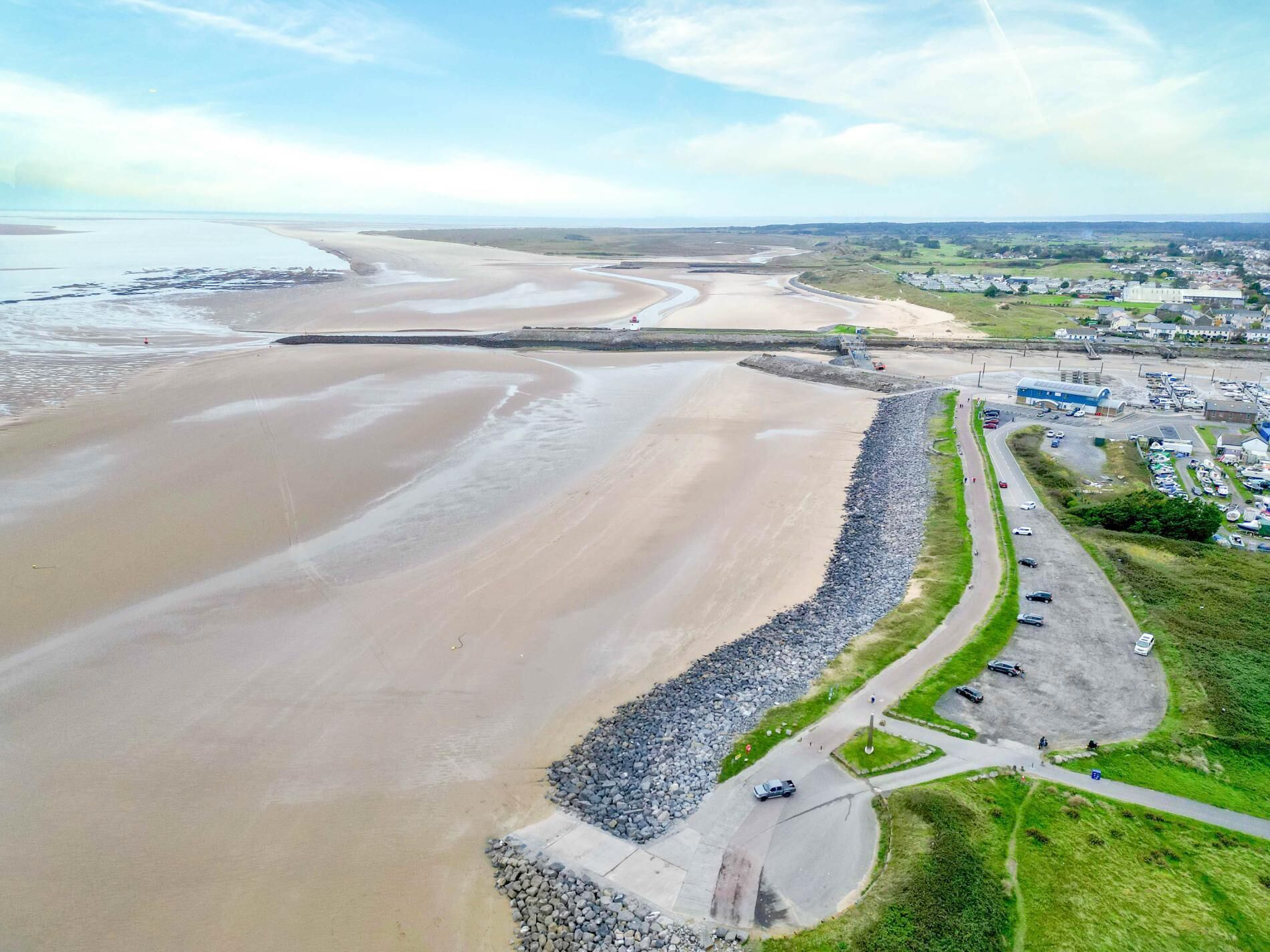 An aerial view of a coastal area with a sandy beach, a rocky breakwater, and a road leading to a small town with buildings and parked cars.