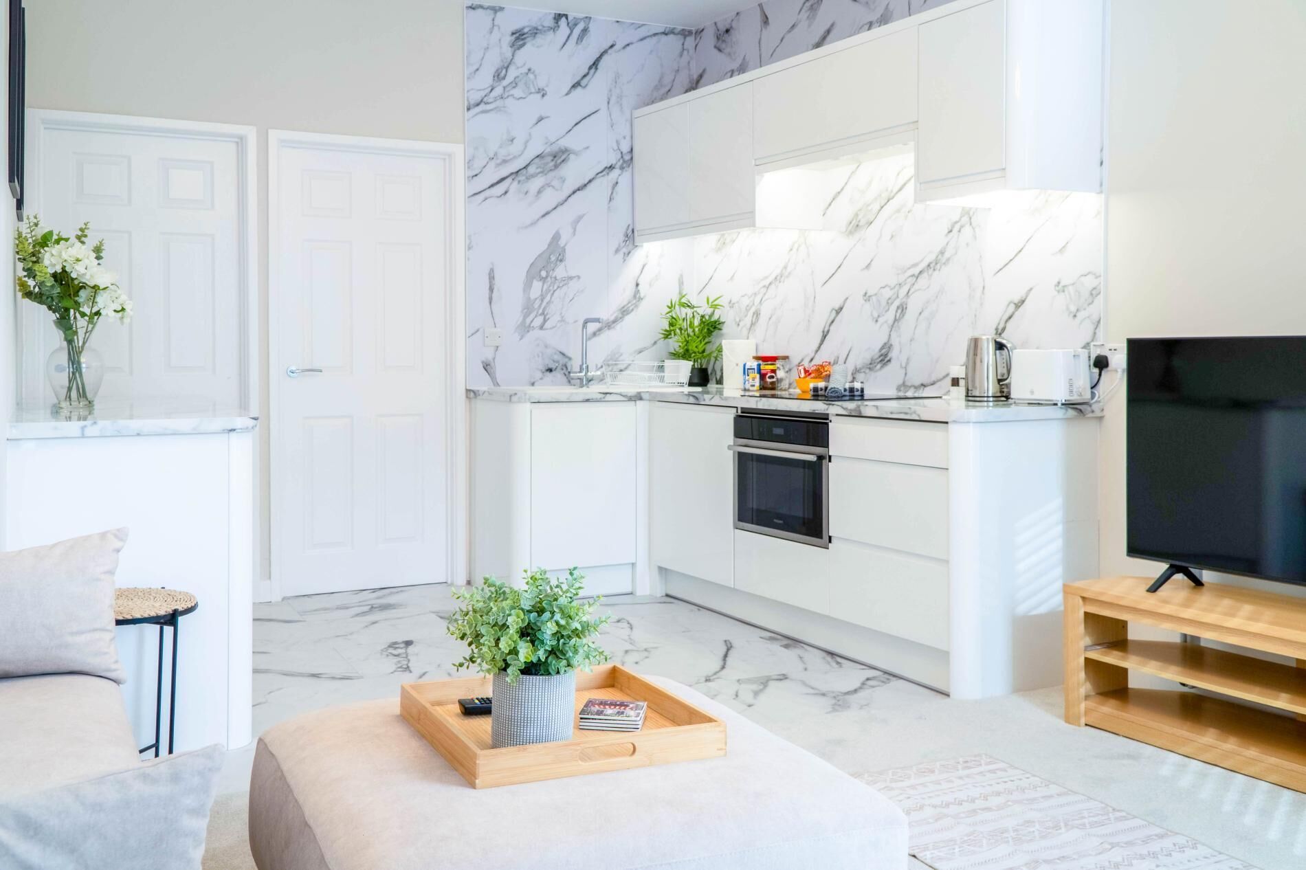 A modern kitchen with white cabinets and marble backsplash, a dining area with a sofa and a coffee table, and a TV on a wooden stand.