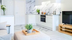 A modern kitchen with white cabinets and marble backsplash, a dining area with a sofa and a coffee table, and a TV on a wooden stand. - Station Road - 1 Bedroom Apt - Burry Port (Burry Port)