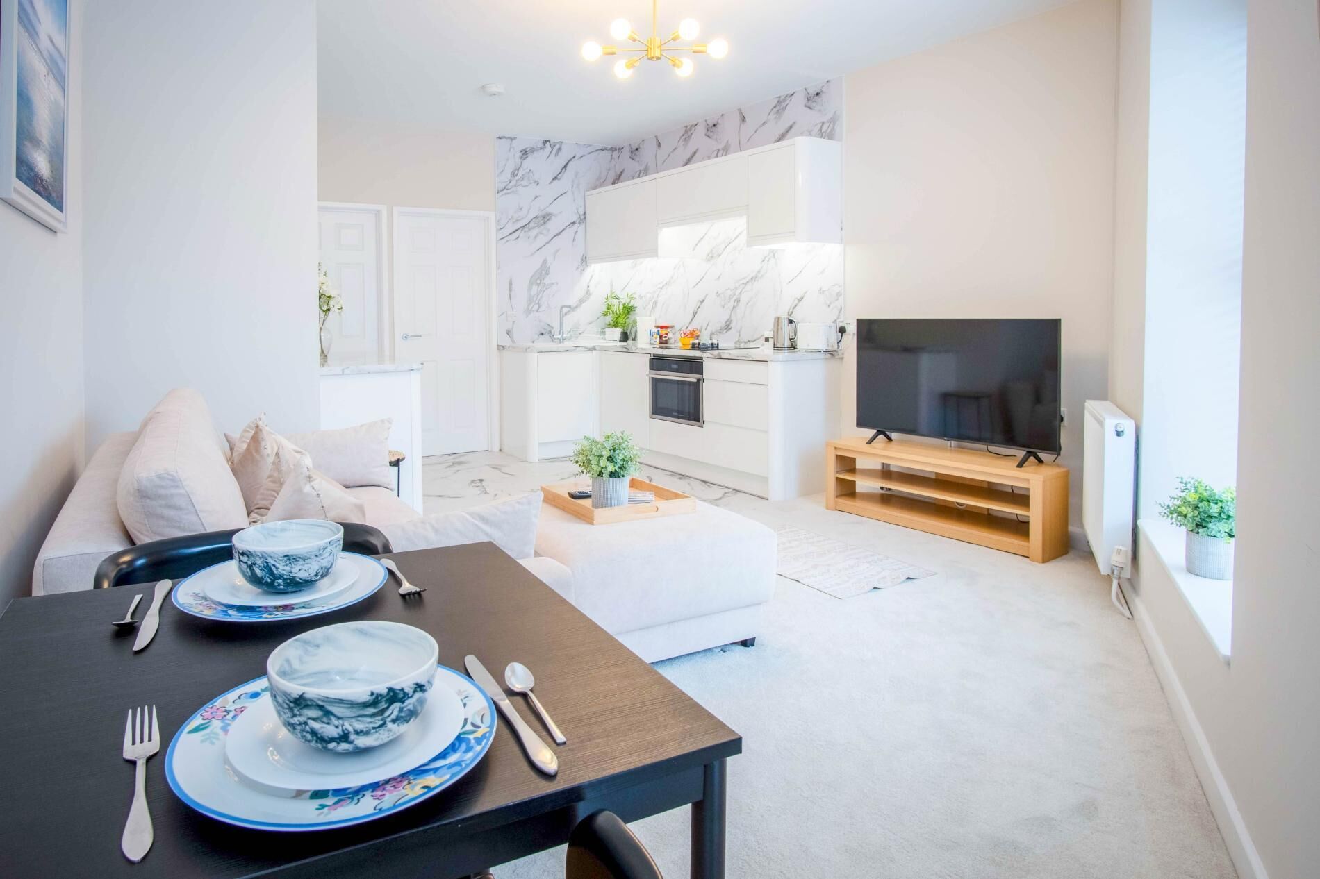 A modern living room with a dining area, a kitchen with marble backsplash, and a flat-screen TV.
