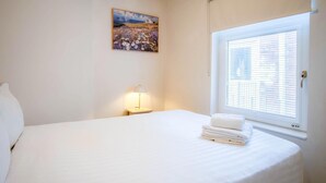 A neatly made bed with white linens and a stack of white towels, a bedside lamp, and a painting on the wall. - Station Road - 1 Bedroom Apt - Burry Port (Burry Port)