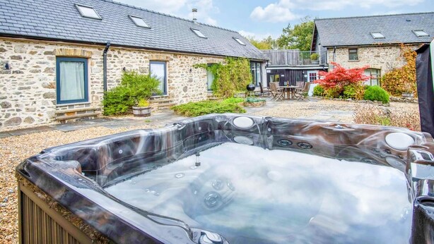 A stone building with a hot tub in the foreground, a patio area, and a small deck with outdoor furniture. - West Barn - 2 Bedroom Cottage - Llandeilo (Llandeilo)
