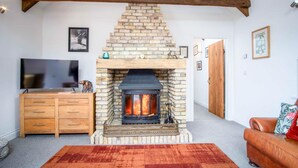 A living room with a stone fireplace, wooden beams, a TV, a wooden cabinet, and a leather sofa. - West Barn - 2 Bedroom Cottage - Llandeilo (Llandeilo)