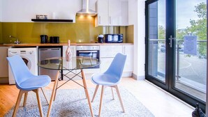 A modern kitchen with a glass dining table, two blue chairs, a coffee machine, and a view of the outside.