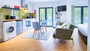 A modern apartment with a kitchen, dining area, and living room. It includes a washing machine, a dining table with chairs, a sofa, and a TV mounted on the wall.