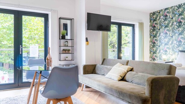 A modern living room with a grey sofa, a glass coffee table, and a grey chair. There is a TV mounted on the wall, a small shelf with plants, and a window with a view of greenery.