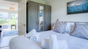 A bedroom with a bed, towels, a wardrobe, and a painting on the wall. - Bayview 72 - 2 Bedroom Chalet - Bayview Oxwich (Swansea)