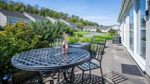 A patio with a round table and chairs, a bottle of wine, and a glass. - Bayview 72 - 2 Bedroom Chalet - Bayview Oxwich (Swansea)