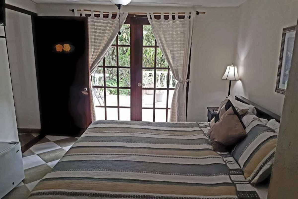 A bedroom with a large bed, a nightstand, a lamp, and a window with curtains.