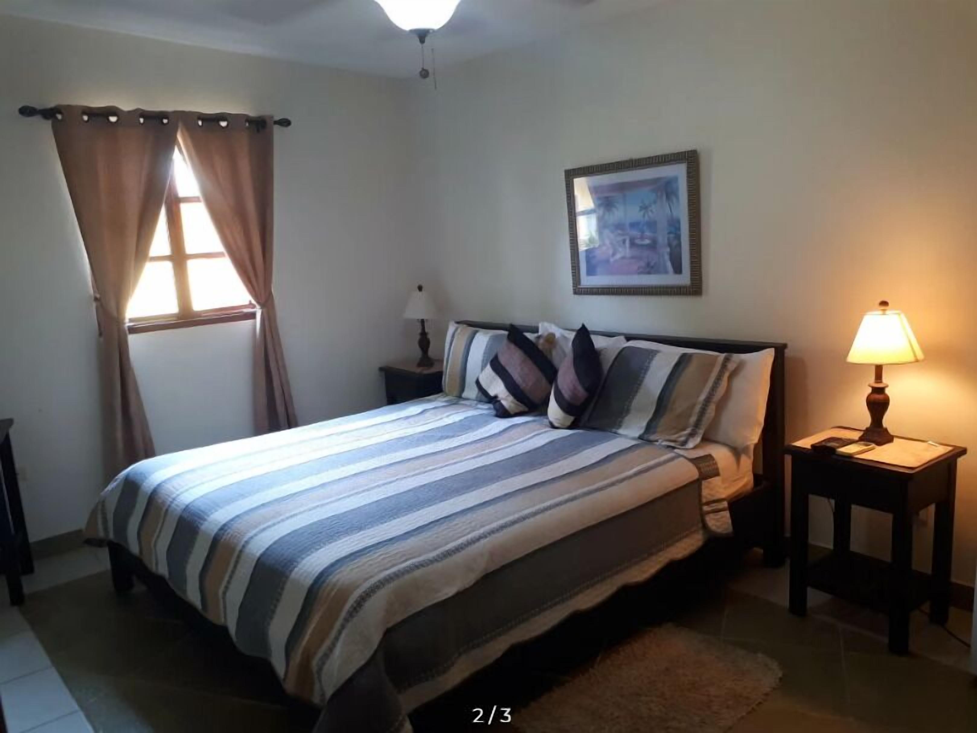 Un dormitorio con cama, dos mesitas de noche, una ventana con cortinas, una lámpara en el techo y un cuadro en la pared.
