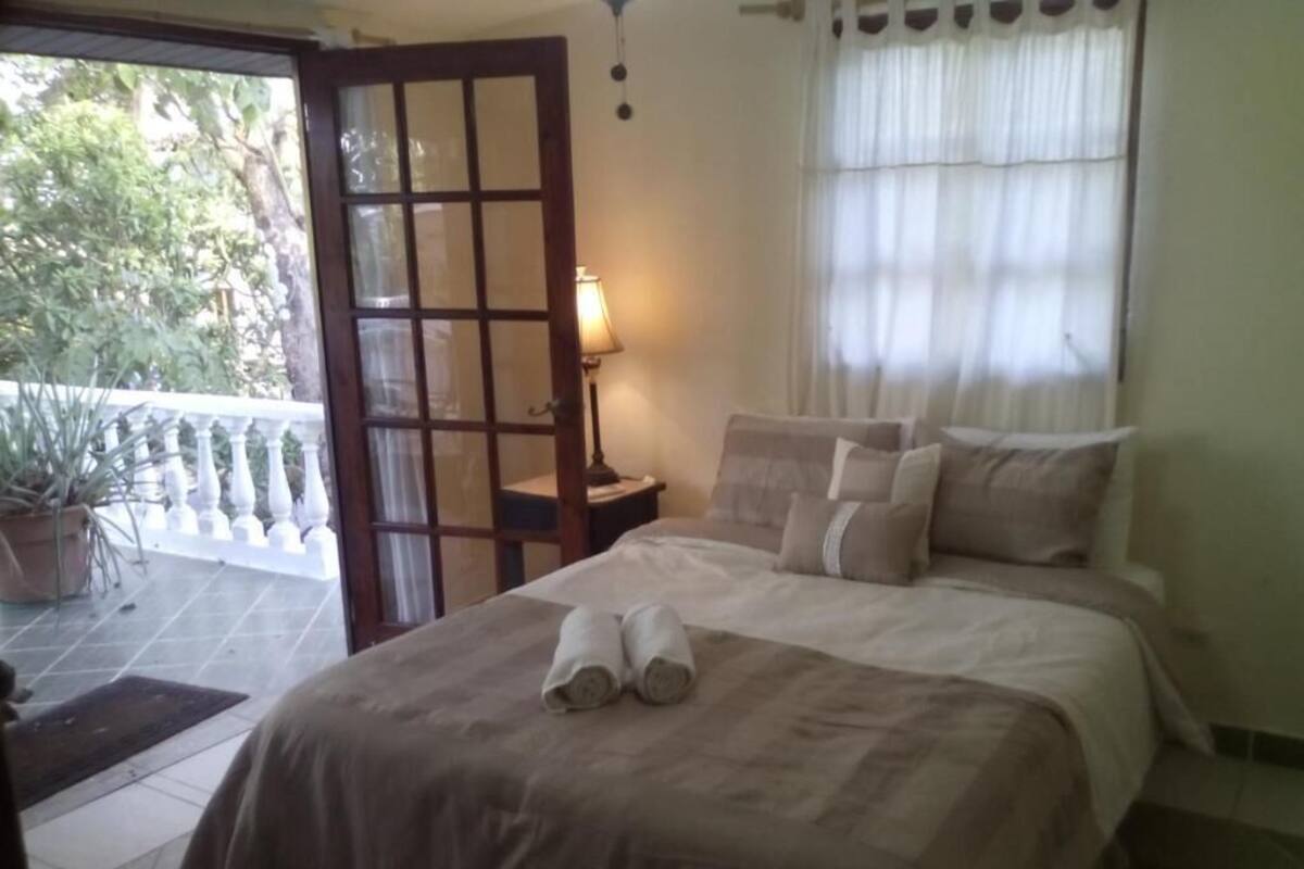 A bedroom with a bed, a nightstand, a window with curtains, and a view of the outdoors.