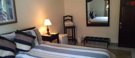 A hotel room with a bed, a bedside lamp, a chair, a small table, a mirror, and a door.