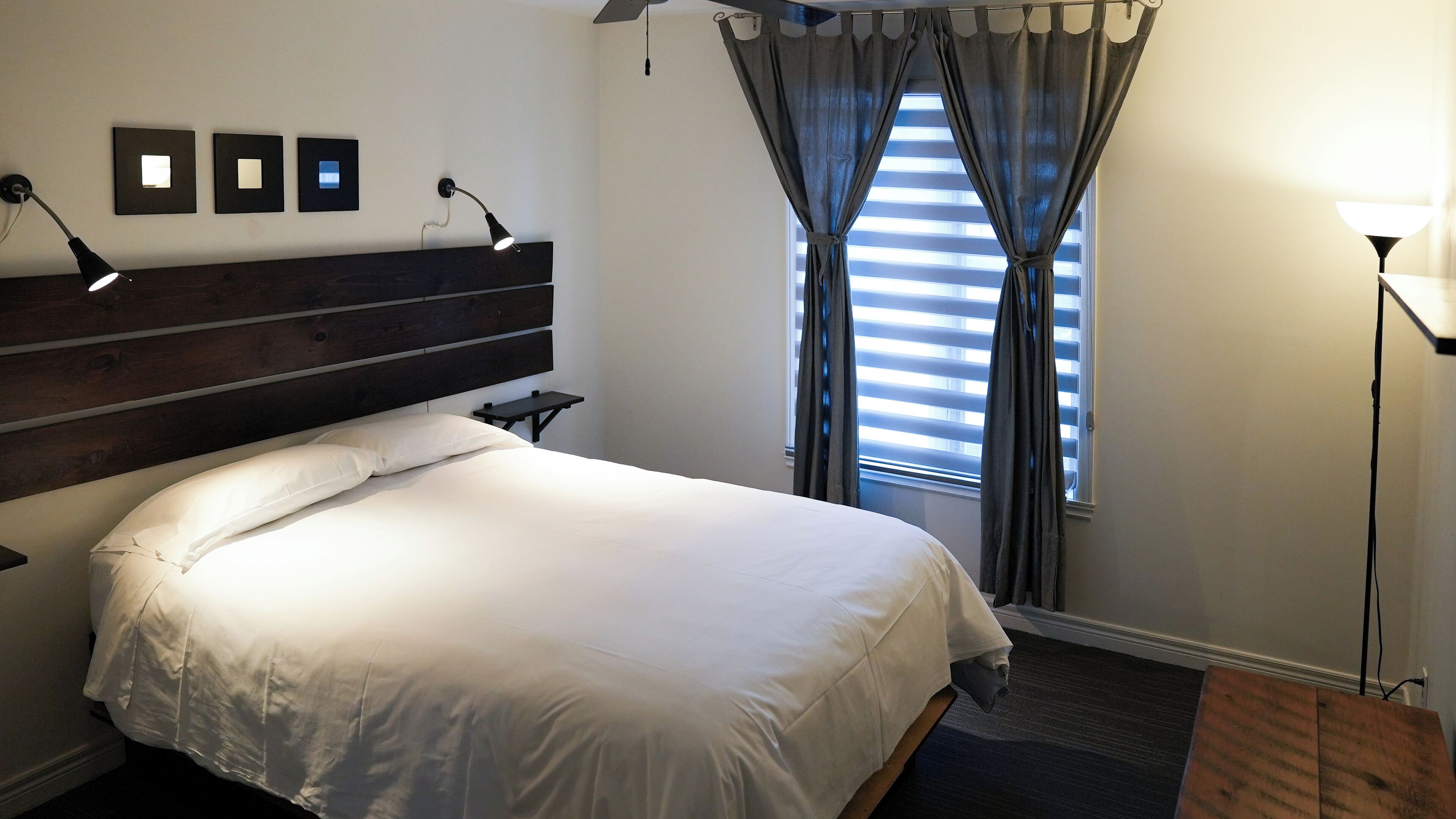 Een opgemaakt bed met witte lakens, een donker houten hoofdbord, een nachtkastje met een lamp, en een raam met gordijnen.