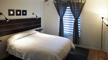 Een opgemaakt bed met witte lakens, een donker houten hoofdbord, een nachtkastje met een lamp, en een raam met gordijnen.