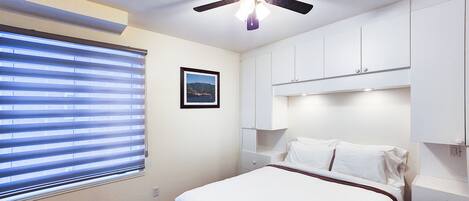 Habitación de hotel con cama, ventilador de techo, ventana con persianas y un cuadro en la pared.