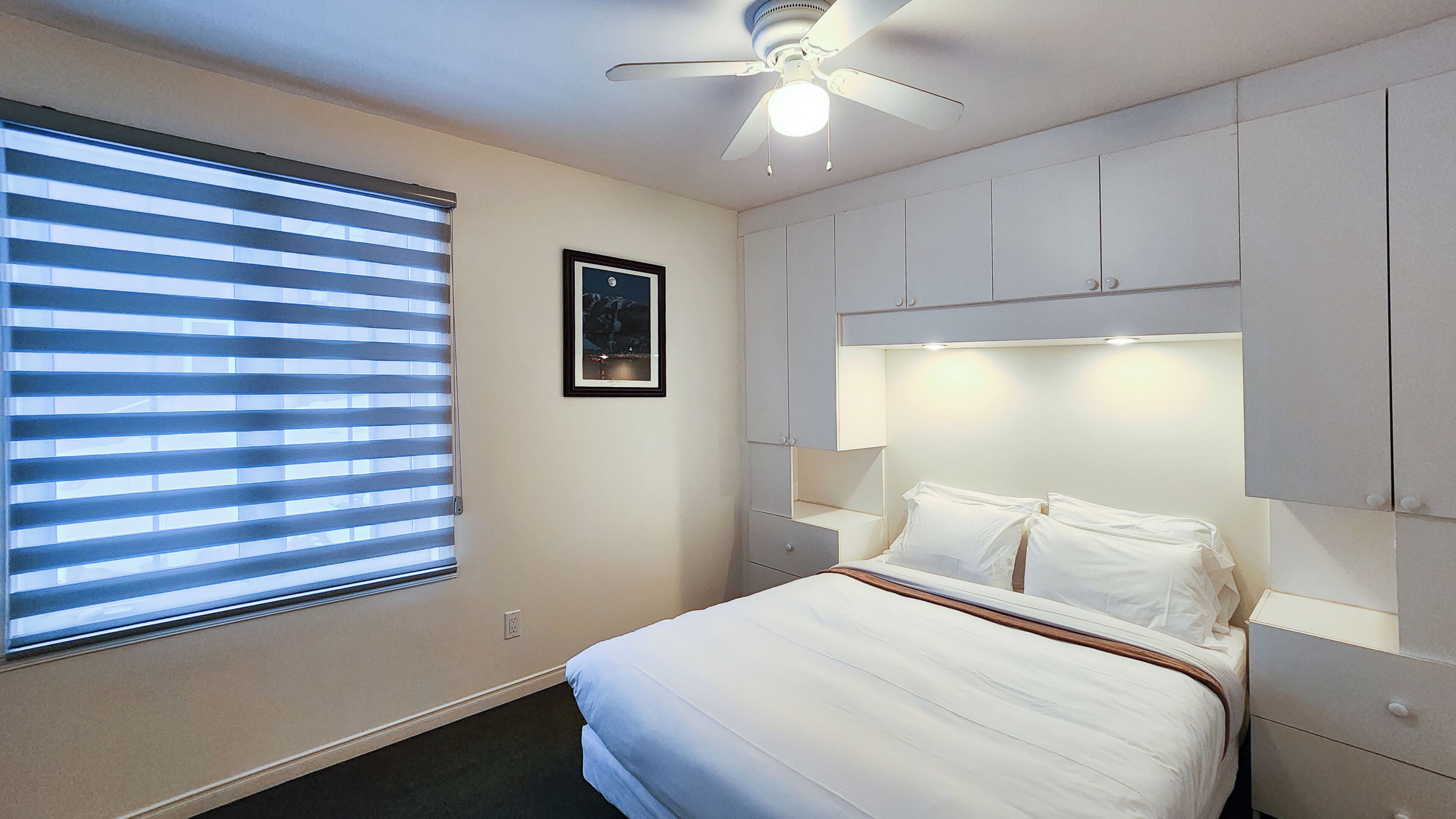 A hotel room with a bed, a ceiling fan, a window with blinds, and a framed picture on the wall.