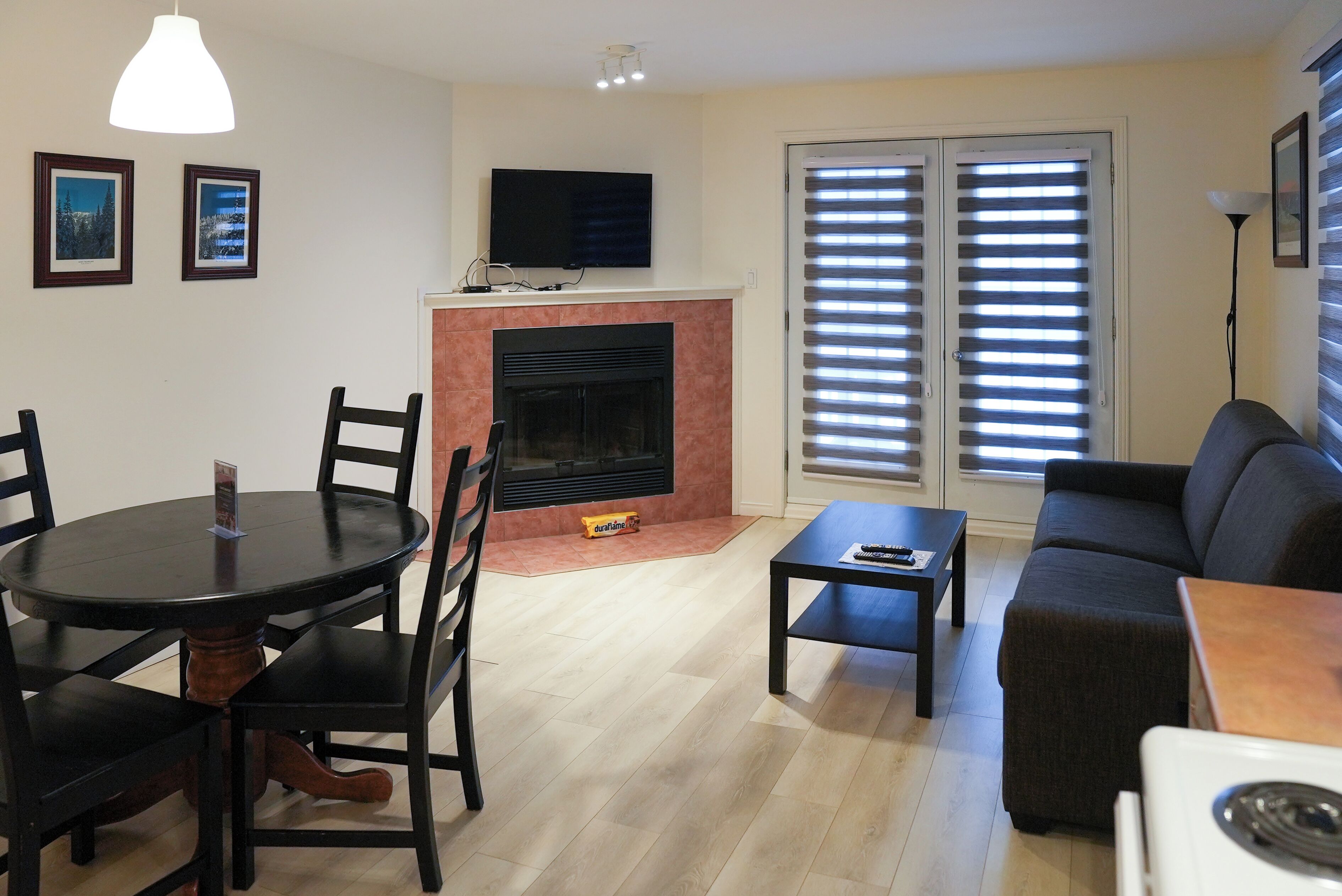 A living room with a fireplace, a television mounted above it, a dining table with chairs, a sofa, and a small table with a lamp.