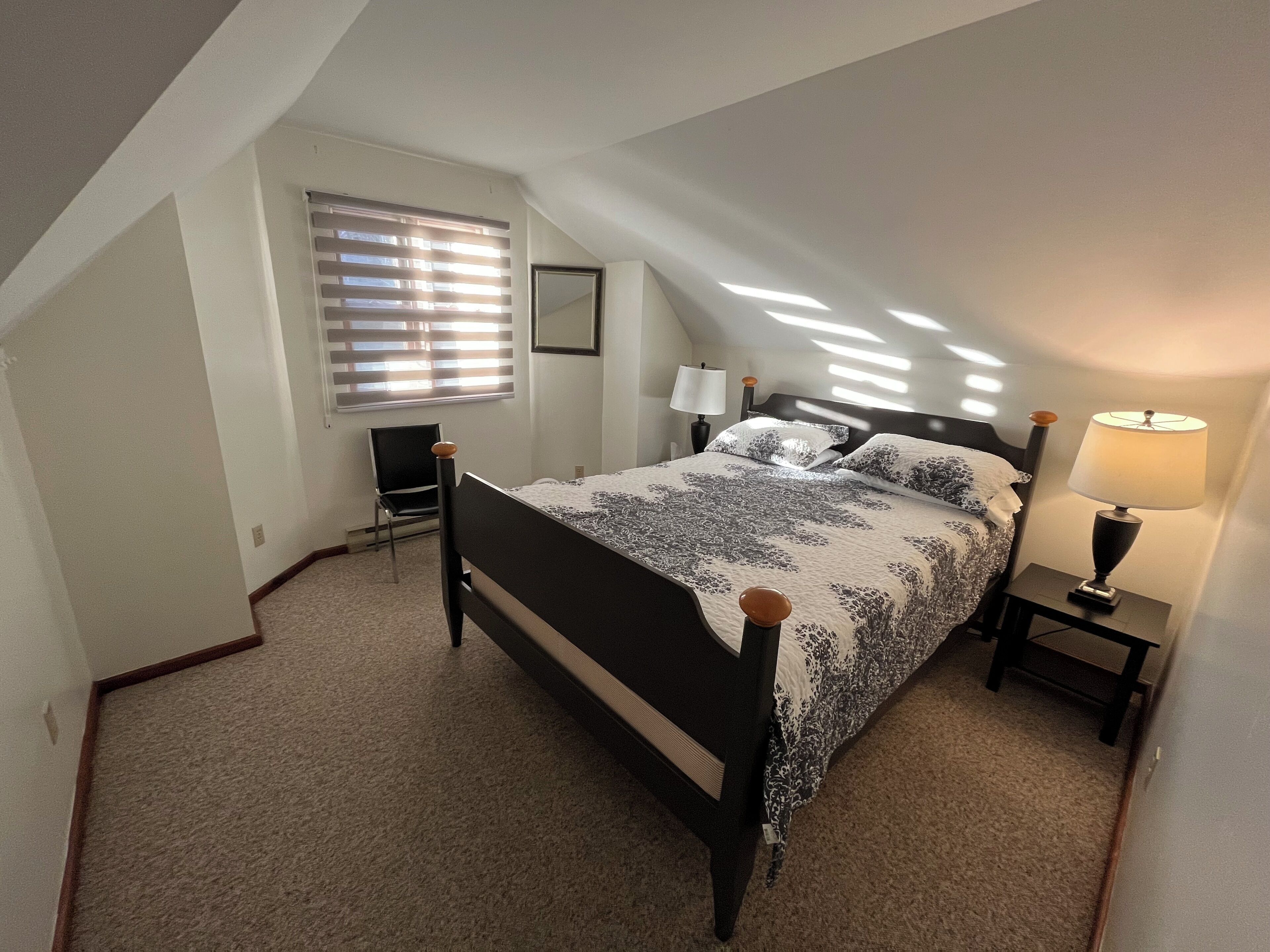 Un dormitorio con techo inclinado, cama con cubrecama estampado, mesita de noche con lámpara, silla y ventana con persianas.