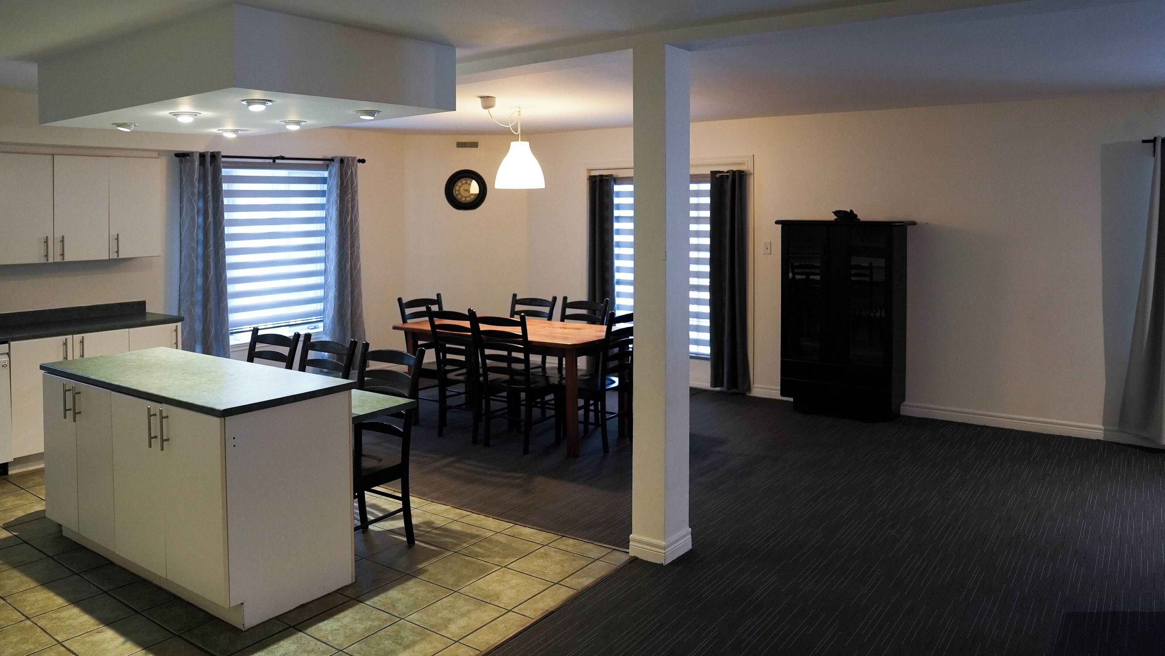 A compact kitchen with a central island, dining area with chairs, a clock on the wall, and a cabinet.