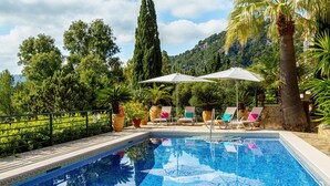 Una piscina rectangular con bordes de azulejos, rodeada de sillones de descanso y sombrillas, con abundante vegetación y una montaña al fondo.