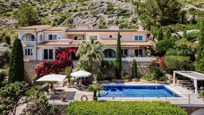Una casa de dos pisos con piscina, rodeada de follaje y una ladera rocosa.