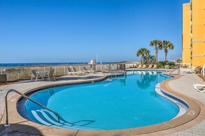 A pool with a clear view of the ocean, surrounded by lounge chairs and a yellow building.