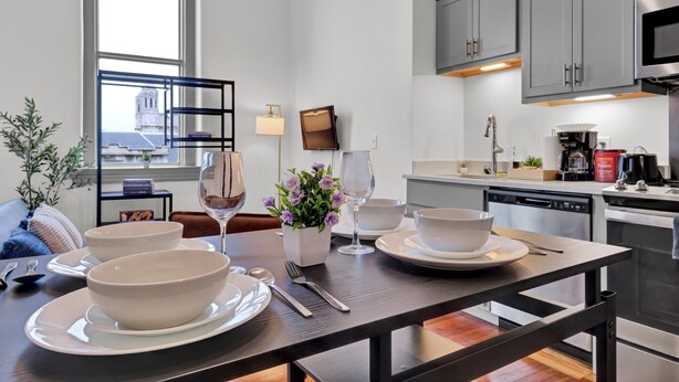 A modern kitchen with grey cabinets, a dining table set with white bowls and plates, a wine glass, and a small potted plant. - Near Hospitals & DT ~ Bright+Parking+W/D+Churchill (Louisville)