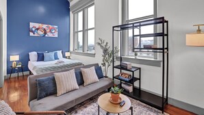 A modern hotel room with a blue accent wall, a bed with blue bedding, a grey sofa with patterned cushions, a small round table with a potted plant, a black bookshelf, and large windows offering a city view. - Near Hospitals & DT ~ Bright+Parking+W/D+Churchill (Louisville)