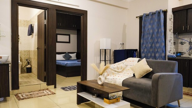 A modern living room with a grey sofa, a coffee table, and a small side table. There is a door leading to a bedroom with a bed and a nightstand. A kitchen area is visible with a sink and cabinets.