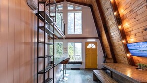 A modern loft-style room with a high ceiling, wooden beams, a mezzanine with a staircase, a large window, a yellow door, and a flat-screen TV mounted on the wall. - 2 Homes-14 Acres+2 Spas +Lake Access+Kayaks (Lakeside)