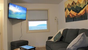 Ein Wohnzimmer mit einem wandmontierten Flachbildfernseher, einem Fenster mit Jalousien, einem grauen Sofa mit Kissen und einem kleinen Tisch mit einem Tablet und einer dekorativen Kugel.