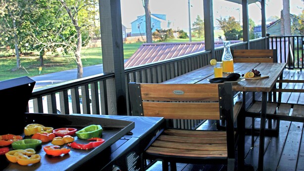Eine Terrasse im Freien mit Holztischen und -stühlen, einem Grill und Gemüse sowie Blick auf ein Haus und Bäume.