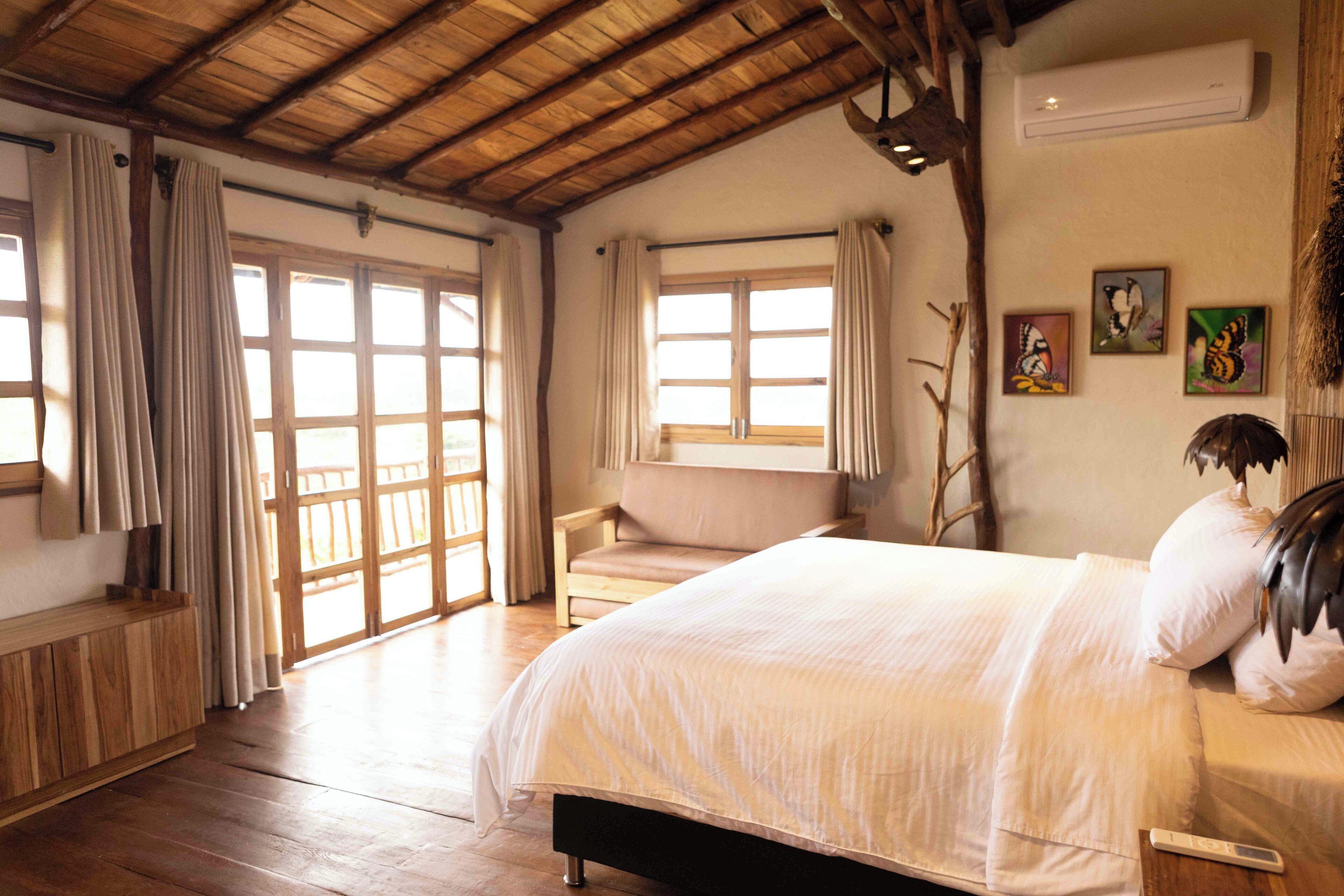 A spacious bedroom with a large bed, wooden flooring, a bench, and a view through large windows.