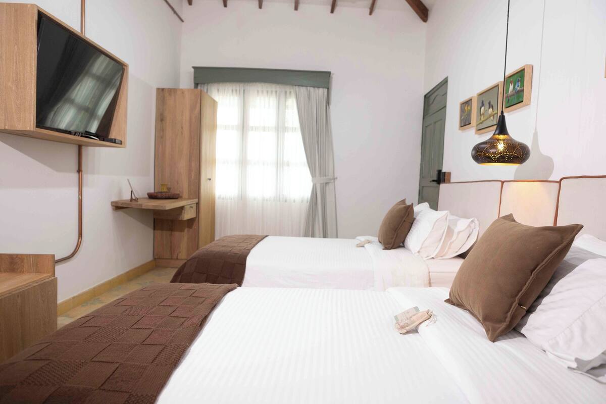 A hotel room with two beds, a TV mounted on the wall, a wooden cabinet, and a window with curtains.