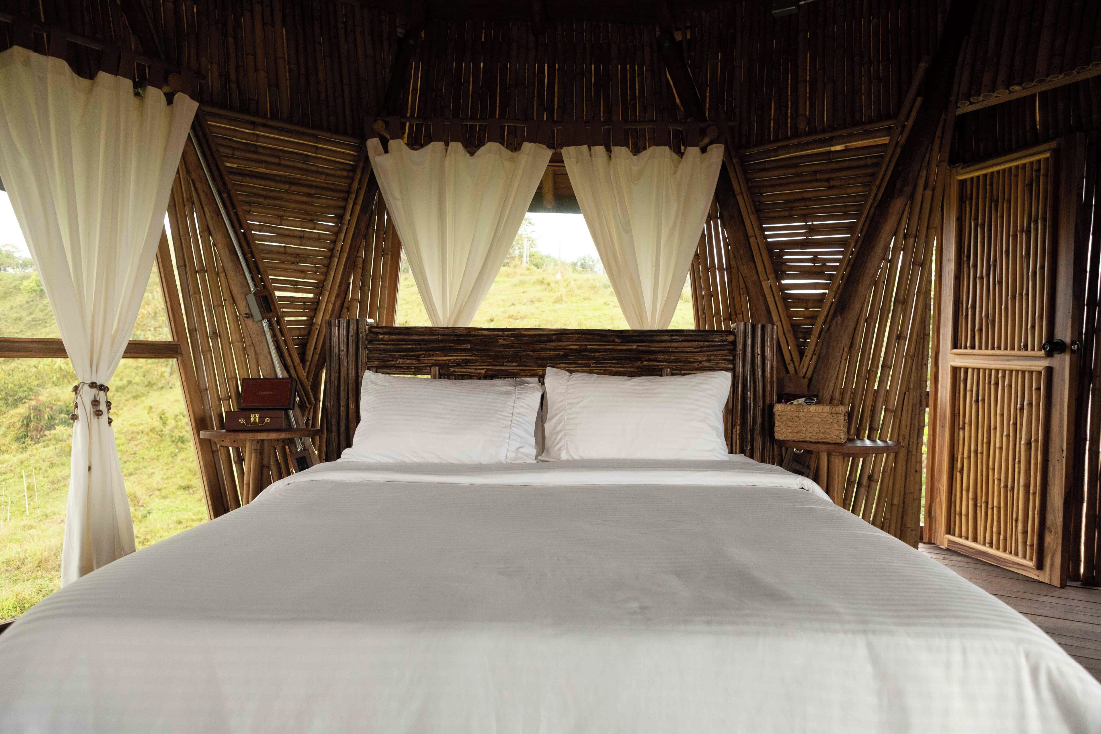 A bed with white linens, a wooden headboard, and white curtains in a room with bamboo walls and a view of the outdoors.