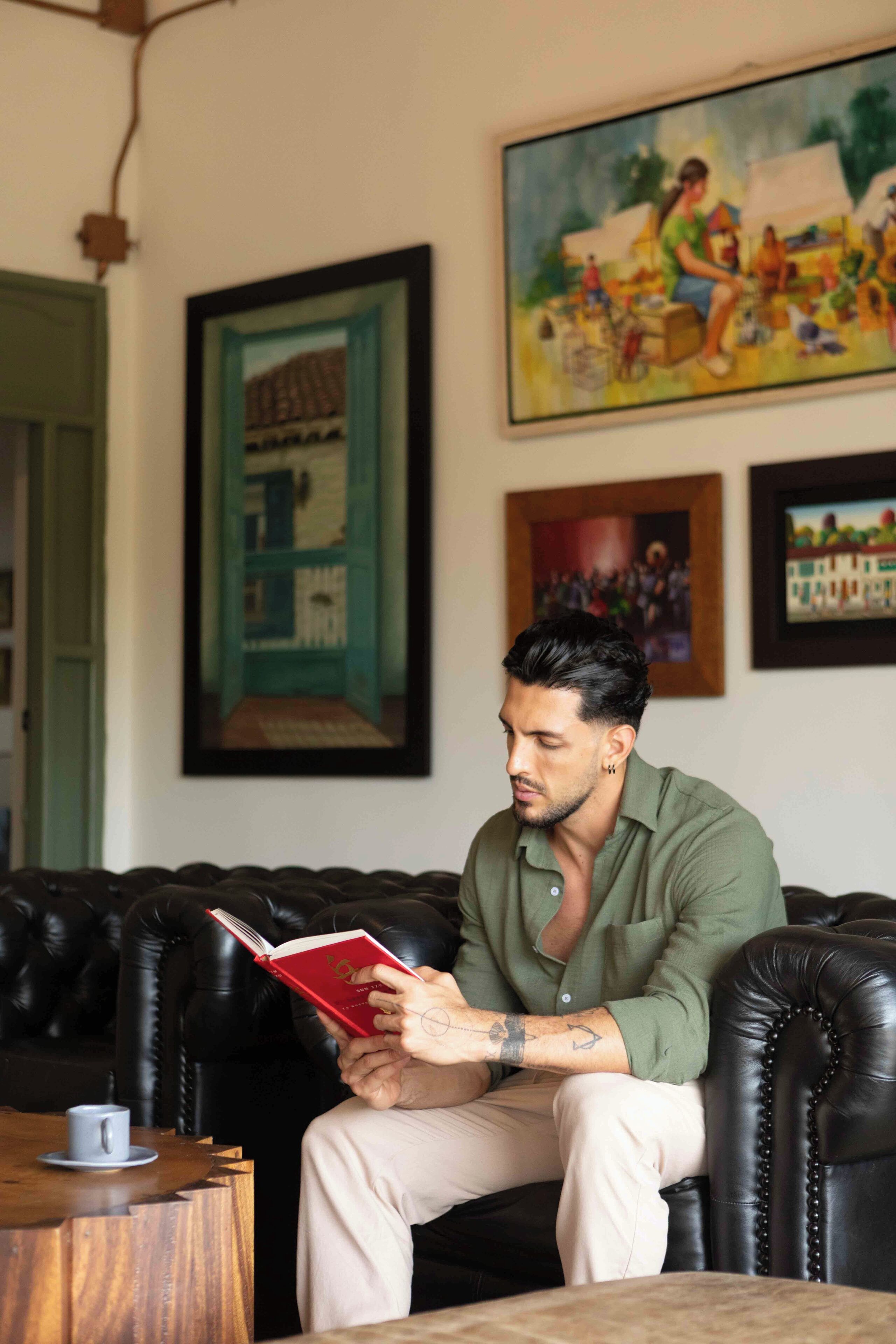 A man sitting on a black leather couch reading a red book. There is a framed painting on the wall behind him.
