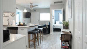 A modern kitchen with a central island, bar stools, a dining area with a table and chairs, and a bedroom with a bed and a nightstand.