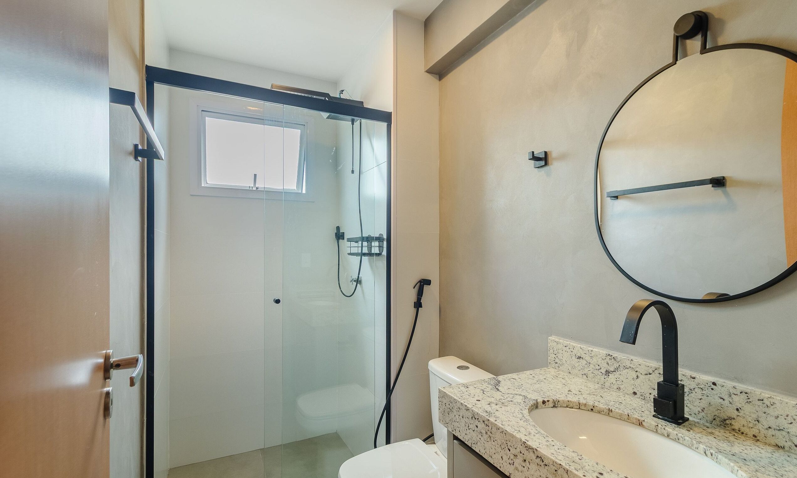 A bathroom with a glass shower enclosure, a toilet, a sink with a granite countertop, and a round mirror.